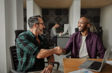 Two smiling businessmen shaking hands across a desk in a modern office setting, with colleagues working in the background.